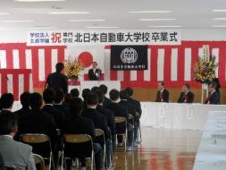 北日本自動車大学校卒業式1。パイプ椅子に座っている卒業生と、国旗と学校旗の前で壇上の市長、来賓の方々と、一人席を立つ卒業生の部屋の後ろからの写真。