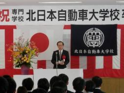 北日本自動車大学校卒業式２。座っている卒業生と、国旗と校旗の前で壇上の市長が壇上に立って話している様子を部屋の後ろから撮った写真。
